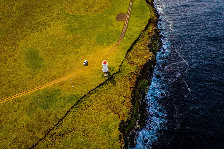 Luftaufnahme eines Leuchtturms auf einer grasbewachsenen Klippe am Meer mit einem Fahrzeug auf einem Weg