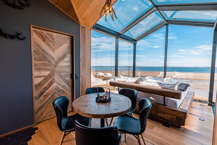 Essbereich mit rundem Holztisch und schwarzen Stühlen neben einer Glasfront mit Blick auf Strand und Meer