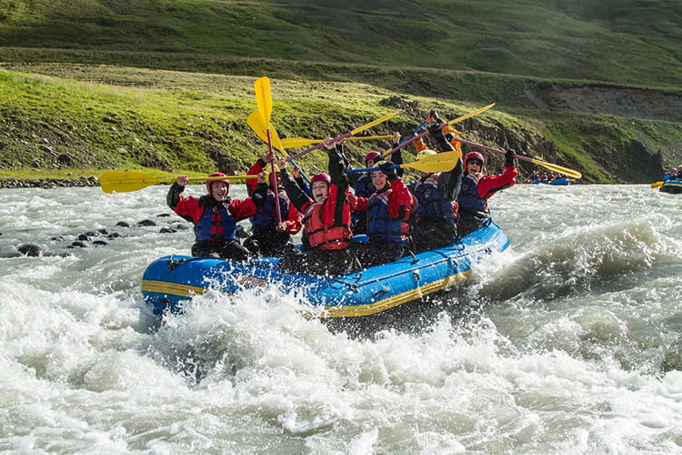 Gruppe von Menschen in roten und blauen Schwimmwesten auf einem blauen Schlauchboot beim Wildwasser-Rafting in einem Fluss mit spritzendem Wasser