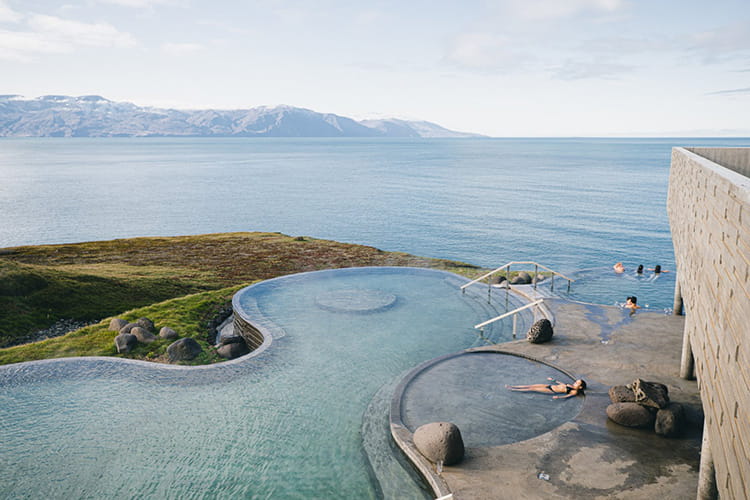 Außenbecken eines Thermalbads mit mehreren Personen, umgeben von Felsen und mit Blick auf ein Meer und entfernte Berge