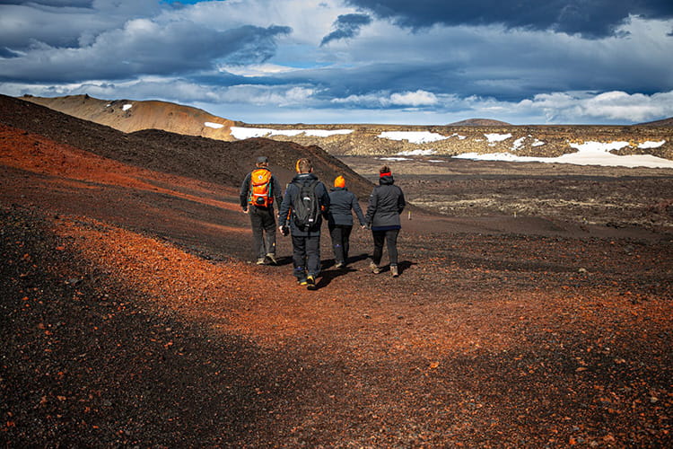 Vier Wanderer mit Rucksäcken gehen über eine karge, vulkanische Landschaft mit vereinzelten Schneeflecken unter bewölktem Himmel