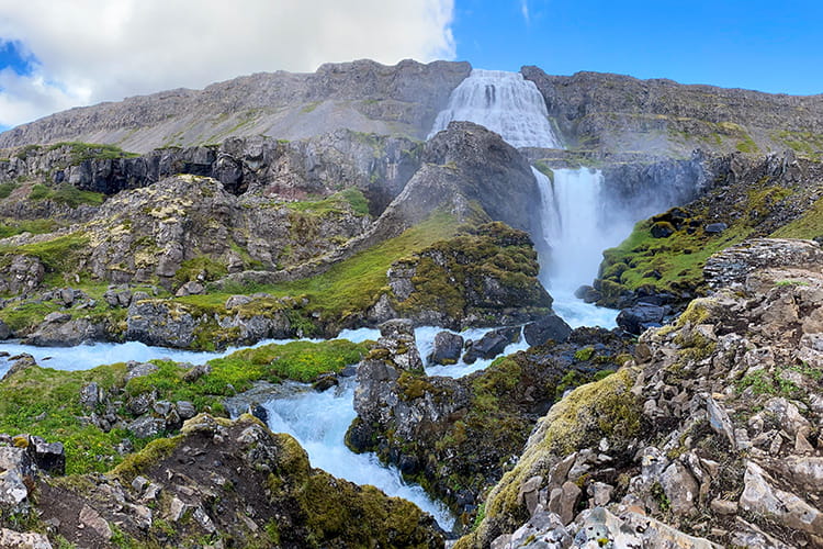 Wasserfall, der in mehreren Stufen über felsige, mit Moos bewachsene Landschaft fließt unter blauem Himmel