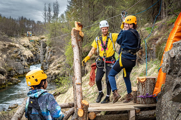 Drei Personen mit Kletterausrüstung und Helmen an einer Zipline über einem Fluss in felsiger Landschaft