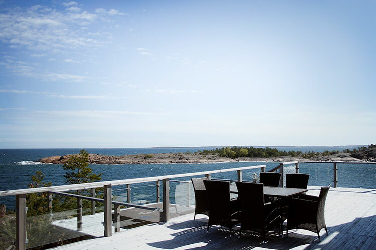 Terrasse mit Tisch und Stühlen aus Rattan vor Meerblick und felsiger Küste unter blauem Himmel