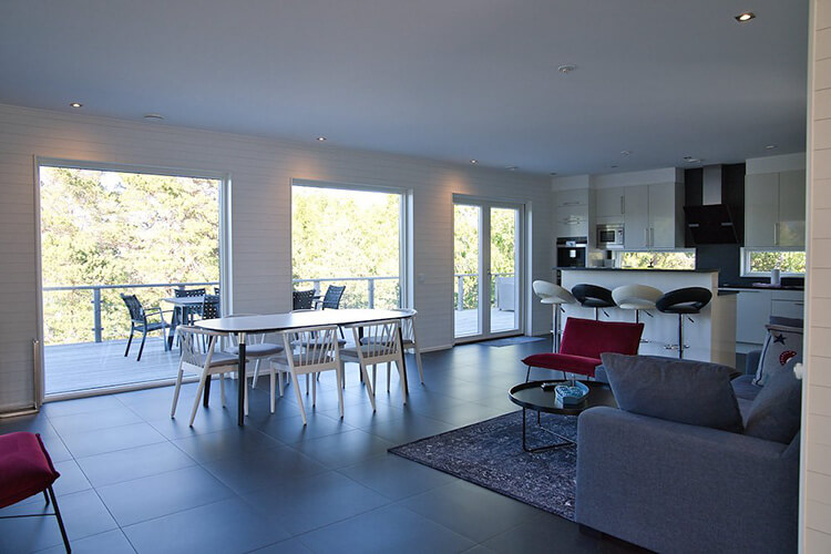 Moderner Wohnraum mit großem Esstisch, grauem Sofa, roten Sesseln und bodentiefen Fenstern mit Blick auf grüne Bäume