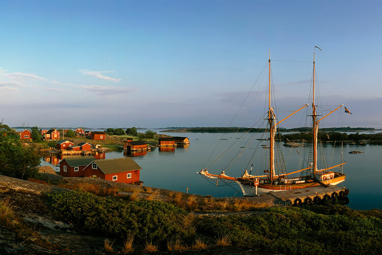 Segelschiff mit zwei Masten liegt an einem Steg vor einer kleinen Siedlung mit roten Holzhäusern an einer ruhigen Küste