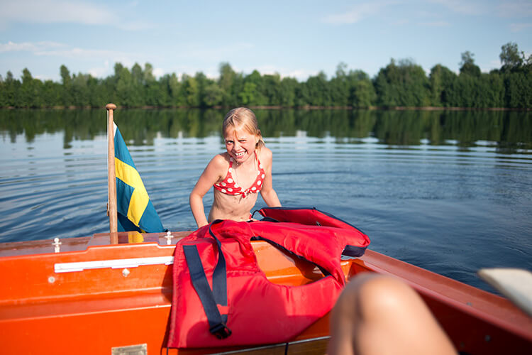 Kind mit rotem Badeanzug sitzt im Boot mit schwedischer Flagge auf ruhigem See