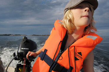 Kind mit orangefarbener Schwimmweste steuert kleines Boot auf offenem Wasser