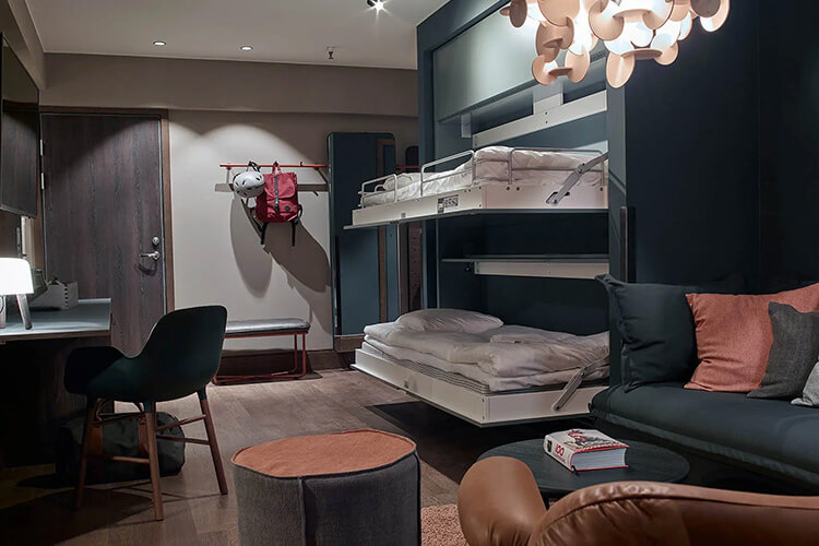 Hotelzimmer mit Etagenbett, Schreibtisch, Stuhl, Sofa, Hocker und Garderobe mit Rucksack an der Wand