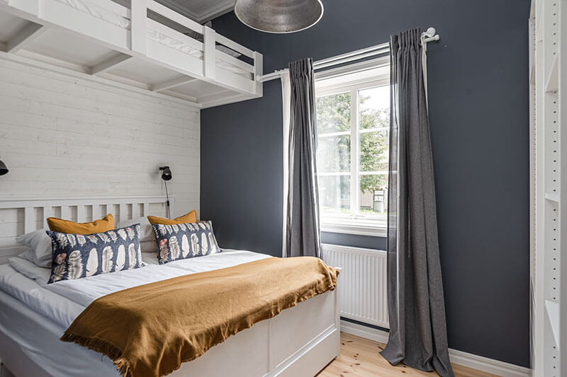 Weißes Doppelbett und ein Hochbett aus Holz in einem Schlafzimmer mit grauen Wänden, Fenster und grauen Vorhängen.