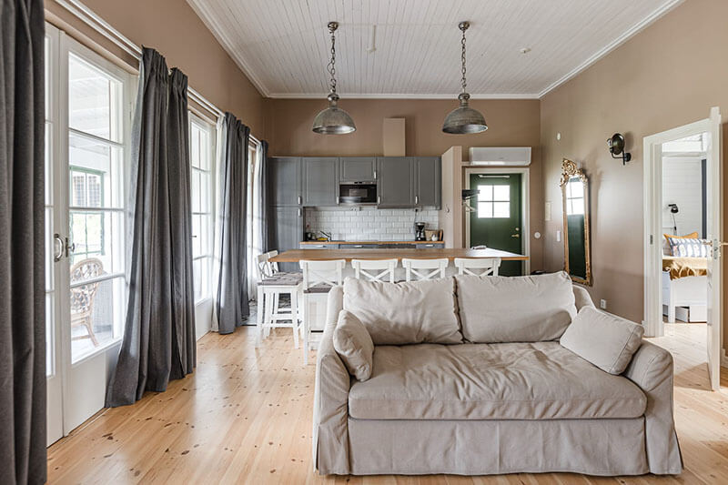 Wohnzimmer und Küche mit einem hellen Sofa im Vordergrund und einer grauen Holzküche mit langem Tisch, Stühlen und Lampen im Hintergrund.