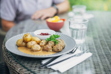 Vor unscharfem Hintergrund mit einer am Tisch sitzenden Person steht ein grauer Teller mit Köttbullar, Kartoffeln und Preiselbeeren auf dem Tisch.