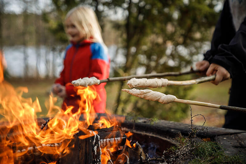 Über einem Lagerfeuer werden Stockbrote gebacken. Im Hintergrund sieht man unscharf ein Kind mit einer roten Jacke und langen blonden Haaren.