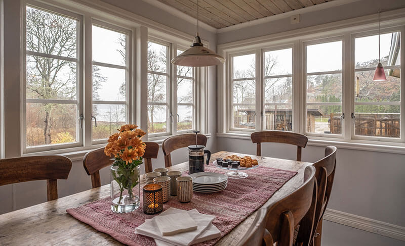 Esszimmer mit Holztisch, sechs Stühlen, Blumenstrauß, Kerzen und Geschirr vor großen Fenstern mit Blick auf Bäume und Wasser.