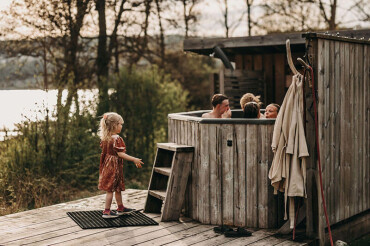 Kind in rotem Kleid steht auf Holzterrasse vor einem hölzernen Hot Tub, in dem mehrere Personen sitzen.