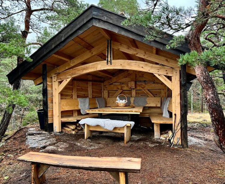 Von Wald umgebene offene Holzhütte mit Sitzbänken und einem Tisch.