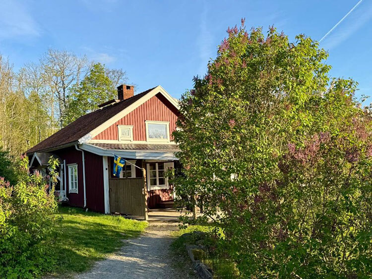 Kleiner Sandweg mit Büschen führt zu einem roten Holzhaus mit einer gelb-blauen Schwedenfahne.