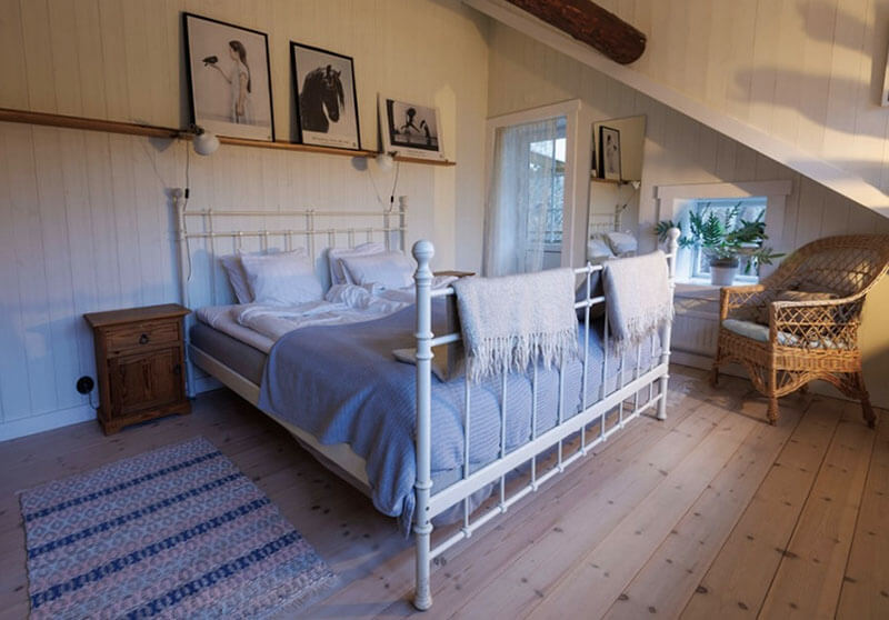 Schlafzimmer mit weißem Metallbett, Holzboden, zwei Nachttischen, drei gerahmten Bildern an der Wand und einem Korbsessel neben einem Fenster.