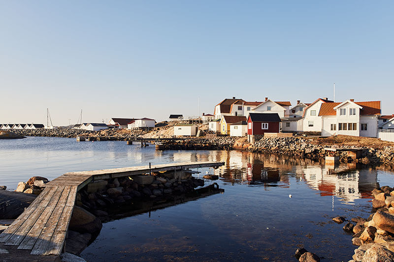 Holzsteg führt über felsige Küste zu kleinen Häusern am ruhigen Wasser unter der Abendsonne.