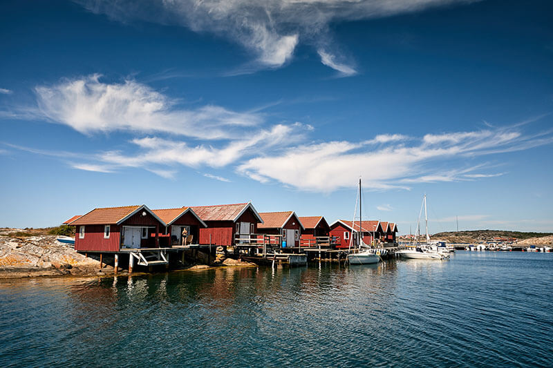 Rote Holzhäuser auf Stelzen am Ufer eines ruhigen Gewässers mit Segelbooten und bewölktem Himmel.
