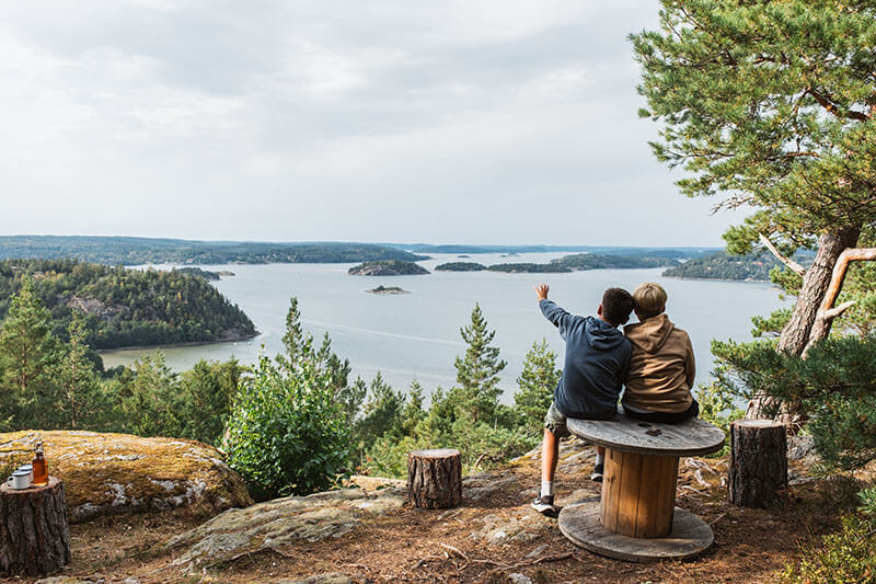 Zwei Kinder sitzen auf einer Holzrolle auf einem Felsen und blicken auf eine Schärenlandschaft mit Wasser und bewaldeten Inseln.