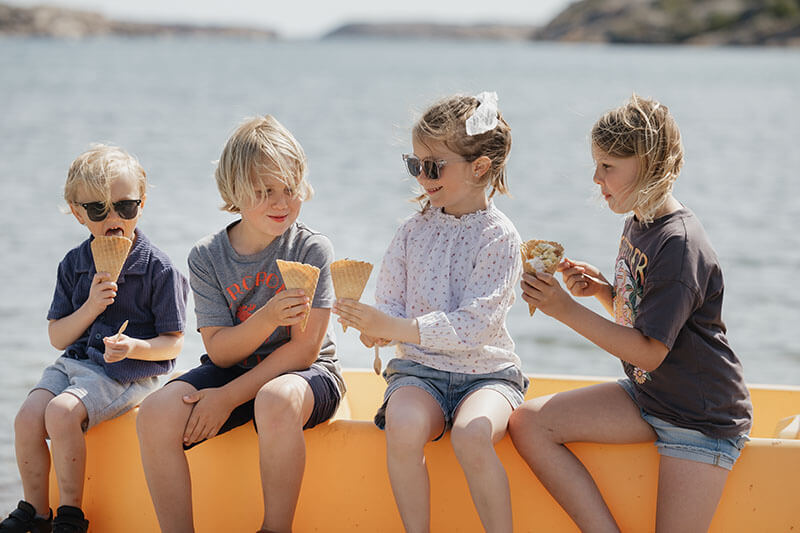 Vier Kinder sitzen nebeneinander auf der Kante eines gelben Bootes am Wasser und essen Eis in Waffelhörnchen.