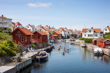 Mehrere Kajakfahrer paddeln durch einen ruhigen Kanal entlang an roten und weißen Holzhäusern.