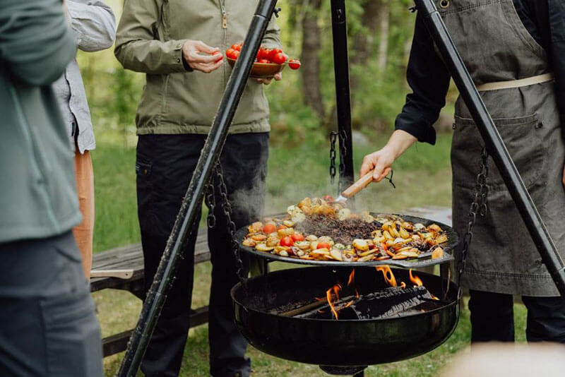 Vier Menschen sind von Wald umgeben und stehen um einen Schwenkgrill, auf dem sie Gemüse und Fleisch zubereiten.