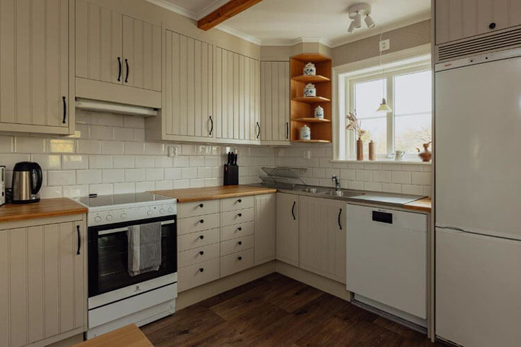 Helle Küche mit weißen Kacheln, Holzarbeitsflächen, Herd, Spüle unter Fenster und offenen Regalen mit Dekorationsgegenständen.