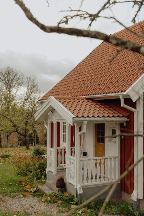 Rotes Holzhaus mit weißem Verandavorbau und brauner Holztür vor herbstlicher Baumkulisse.