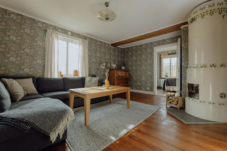 Wohnzimmer mit Kachelofen, Sofa, Tisch, Kommode, Lampen, Holzboden und Blümchentapete.