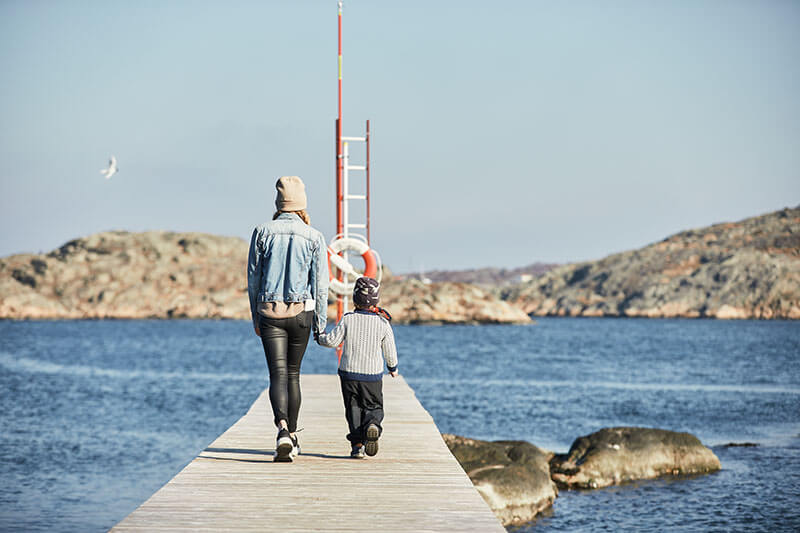 Eine Frau und ein kind laufen auf einem Holzsteg Richtung Meer. Im Hintergrund ist verschwommen die felsige Küstenlandschaft zu sehen.