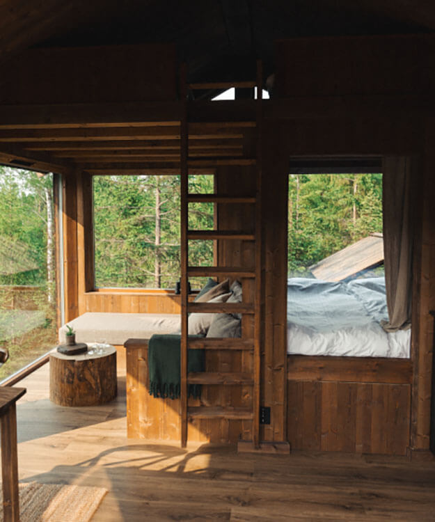 Holzinnenraum mit Fensterfront zum Wald, Holzleiter zu einer Schlafkoje und Bett mit Decke und Kissen.