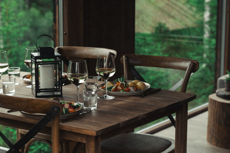 Holztisch mit gedeckten Tellern, Gläsern und einer Laterne vor großen Fenstern mit Blick auf Wald.