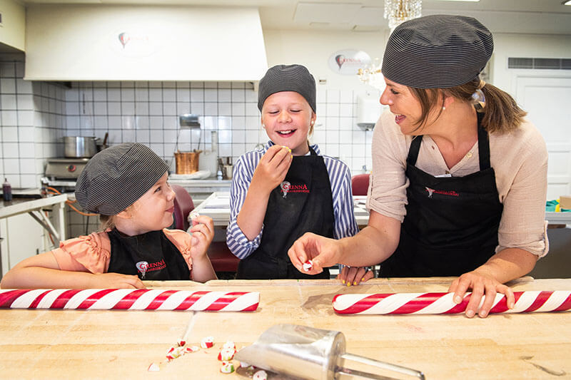 Gemeinsam mit einer Frau stehen zwei Kinder in einer Küche, in der rot-weiß gestreifte Zuckerstangen hergestellt werden.