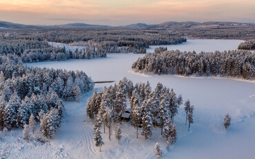 Verschneite Waldlandschaft mit zugefrorenem See und einzelnen Holzhäusern in Abendstimmung