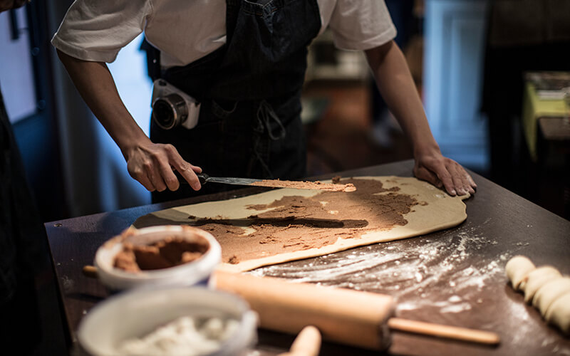 Eine Person, von der man nur den Oberkörper sieht, streicht eine braune Masse auf einen Teig.