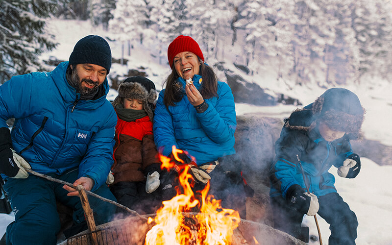 Eine Familie sitz in einer verschneiten Landschaft am Lagerfeuer.