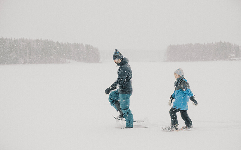 Zwei Personen in Winterkleidung mit Schneeschuhen gehen durch eine verschneite Landschaft mit Bäumen im Hintergrund