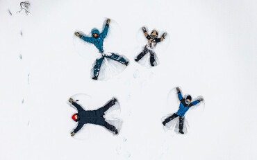 Vier Personen liegen mit ausgestreckten Armen und Beinen im Schnee und machen Schneeengel aus der Vogelperspektive