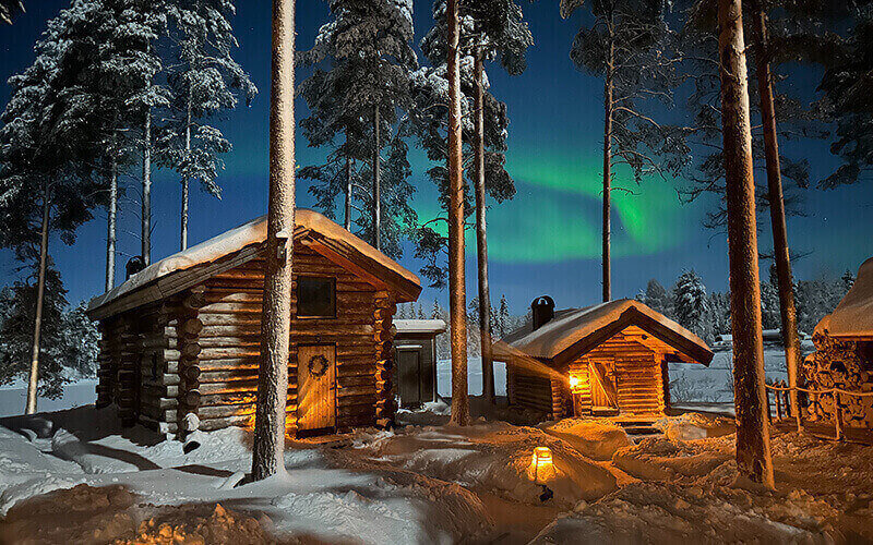Zwei Holzhäuser bei Nacht zwischen Bäumen. Am Himmel sind Nordlichter.