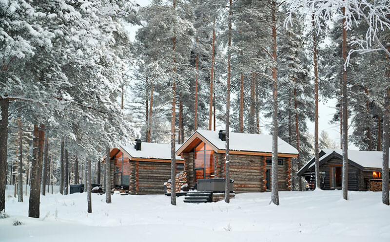 Drei Holzhäuser, in denen Licht brennt, zwischen Bäumen in einer Winterlandschaft.
