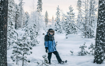 Ein Kind steht in einem blauen Schneeanzug in einer Winterlandschaft und schaut nach unten.