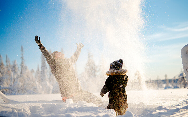 Eine Person und ein kleines Kind im Schnee. Die große Person wirft Schnee in die Luft.