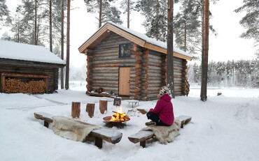 Eine Person sitz auf einem Fell auf einer bank am Lagerfeuer. Eine Blockhütte und ein kleineres Gebäude stehen im Hintergrund.