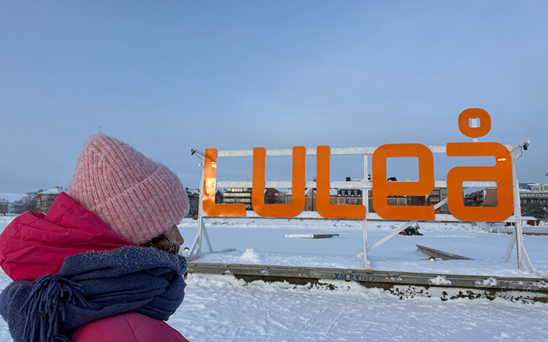 Eine Person mit einer pinken Mütze und pinker Jacke steht vor einem orangenen Schild, das das Wort Luleå bildet.
