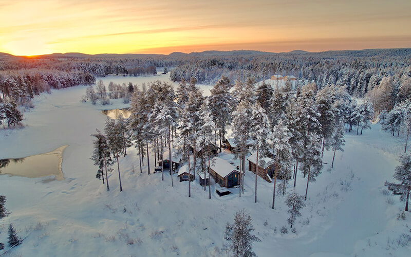 Eine Häuseransammlung aus der Vogelperspektive in einer weiten Landschaft. Es liegt Schnee und die Sonne geht unter.