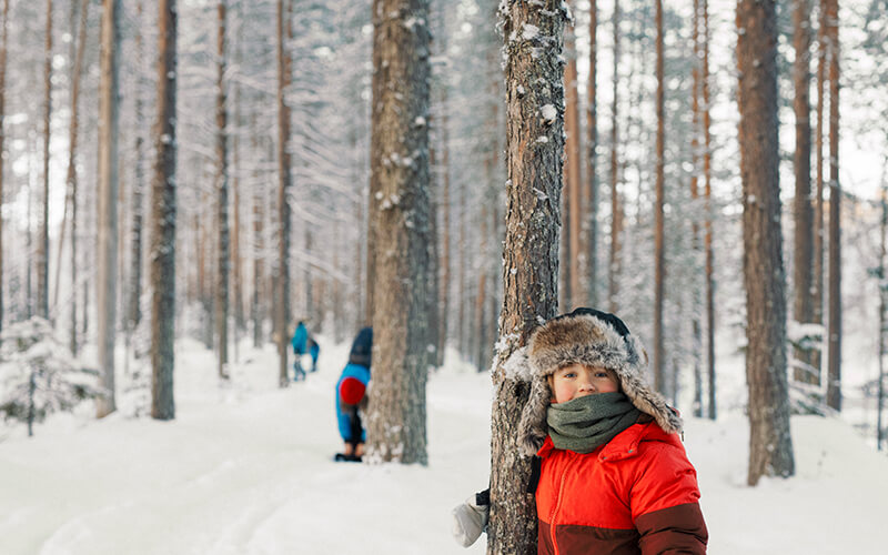Ein kleines Kind steht in einem roten Schneeanzug und einer Pudelmütze neben einem Baum in einem verschneiten Wald.