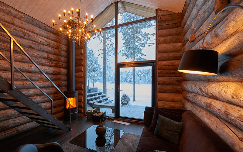 Ein Holzhaus mit großem Glasfenster. Es brennt ein kain in der Ecke, draußen liegt Schnee.