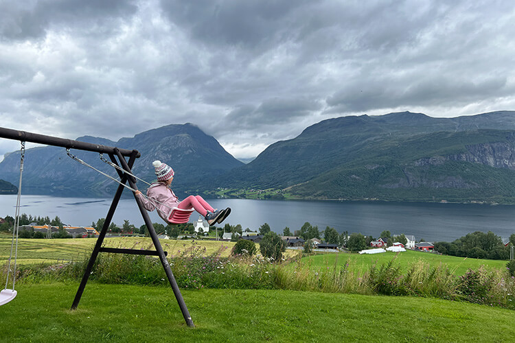 Kind auf einer Schaukel, im Hintergrund Fjordlandschaft und Berge.
